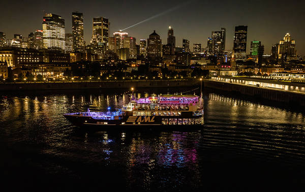 A stunning nighttime view of an illuminated cruise ship sailing on a river, with a sparkling city skyline in the background featuring skyscrapers and urban lights.