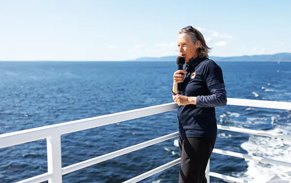 A naturalist holds a microphone and gives a presentation on the deck of a boat, with a clear view of the blue ocean in the background. She is wearing a navy-blue sweater and stands near the white railing under a sunny, clear sky, sharing information about the surrounding nature.