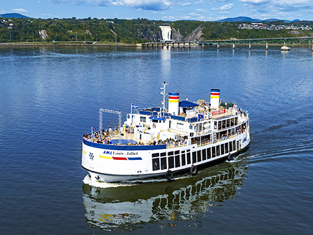 The ship "Louis Jolliet" from AML cruises sails on a calm river during the day. Passengers stand on the decks, enjoying the surrounding scenery, including a waterfall and green hills under a blue sky.