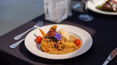 A gourmet plate featuring a chicken leg with sauce, garnished with vegetables, mashed potatoes, and edible flowers, served on an elegantly set table with a dark tablecloth.