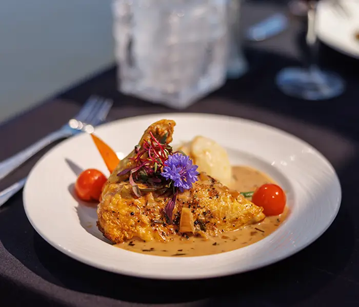 A gourmet plate featuring a chicken leg with sauce, garnished with vegetables, mashed potatoes, and edible flowers, served on an elegantly set table with a dark tablecloth.