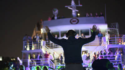A DJ performs at a nighttime party on a boat, with colorful lights and a crowd dancing across multiple levels of the vessel.