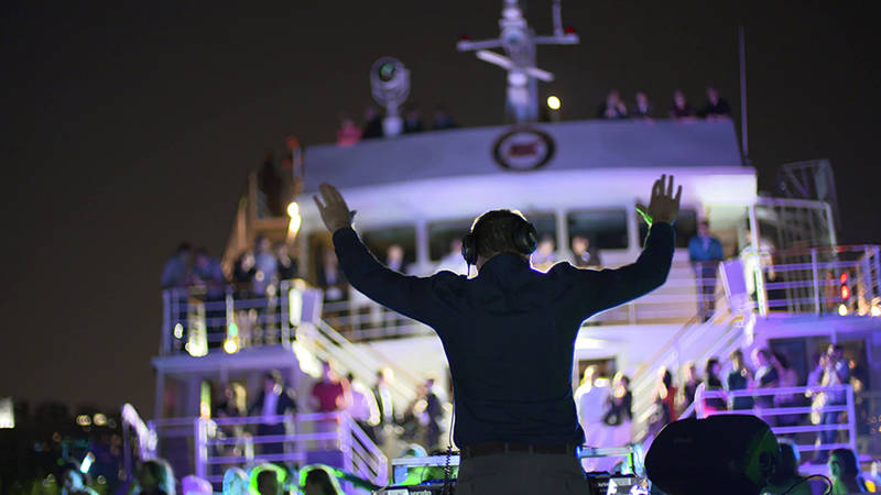 A DJ performs at a nighttime party on a boat, with colorful lights and a crowd dancing across multiple levels of the vessel.