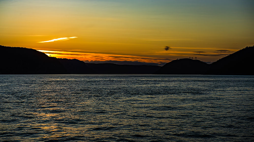 A vibrant sunset lights up a calm body of water, surrounded by dark hills. The sky showcases shades of yellow, orange, and deep blue, subtly reflected on the surface of the water.