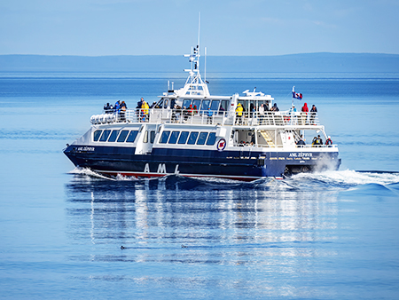 The ship "AML Zéphyr" sails on calm waters with passengers onboard, enjoying a sunny day. The boat is blue with white details and displays a flag. The water slightly reflects the vessel.