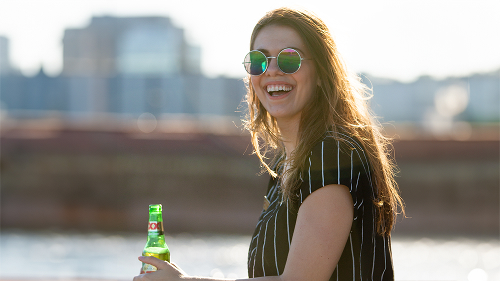 A smiling woman wearing round sunglasses and a striped shirt, holding a green beer bottle, with a river and blurred buildings in the background.
