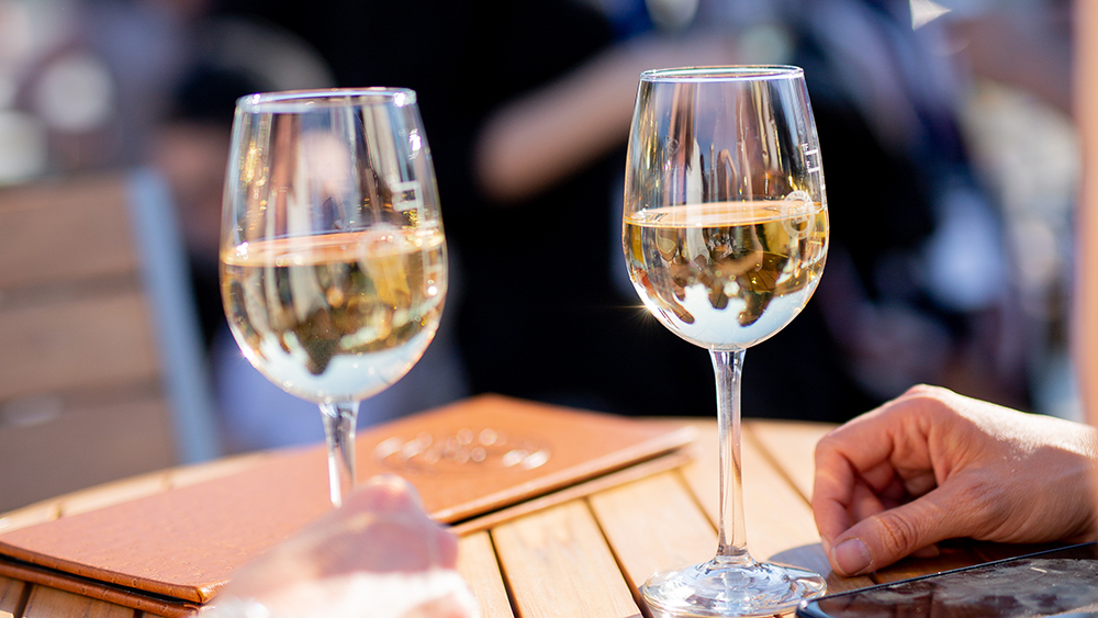 Two glasses of white wine on a wooden table on a sunny day, with visible hands and a blurred background of a lively outdoor setting.