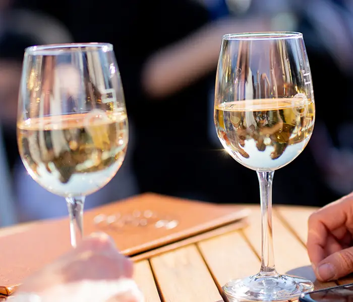 Deux verres de vin blanc posés sur une table en bois lors d'une journée ensoleillée, avec des mains visibles et un arrière-plan flou d'une scène extérieure animée.