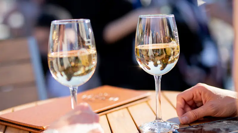 Deux verres de vin blanc posés sur une table en bois lors d'une journée ensoleillée, avec des mains visibles et un arrière-plan flou d'une scène extérieure animée.