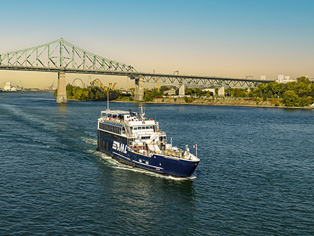 A blue cruise ship sails on a river on a sunny day, with the Jacques-Cartier Bridge and trees in the background.