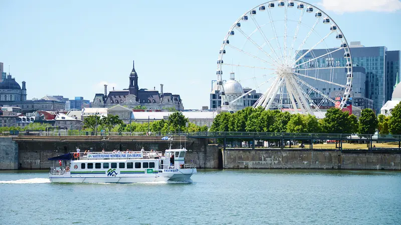 Une vue d'un bateau naviguant sur le fleuve devant une grande roue emblématique, avec des bâtiments historiques et modernes en arrière-plan sous un ciel bleu.