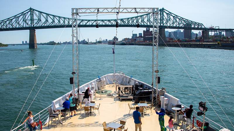 View of the front outdoor terrace of the AML Cavalier Maxim boat, where customers are enjoying the view of the Saint Lawrence River, the Jacques Cartier Bridge, and the city of Montreal in the background.