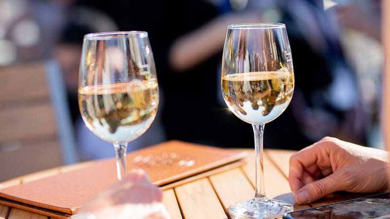 Two glasses of white wine placed on a wooden table, with leather menus and a hand visible in the foreground. The background is blurred, suggesting a bright and sociable outdoor atmosphere.