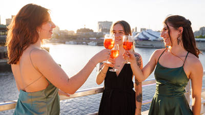 Three elegantly dressed women toast with orange cocktails on a boat deck, with a view of the water and the city at sunset.