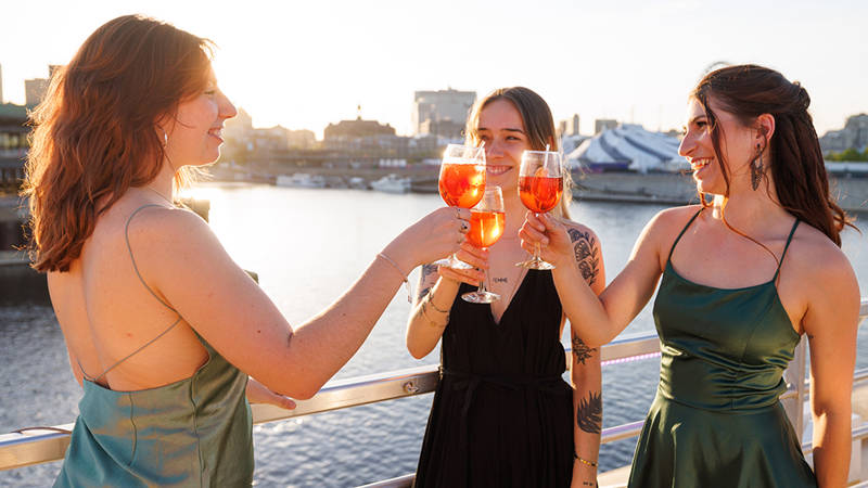 Three elegantly dressed women toast with orange cocktails on a boat deck, with a view of the water and the city at sunset.