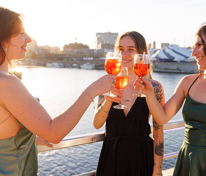 Three elegantly dressed women toast with orange cocktails on a boat deck, with a view of the water and the city at sunset.