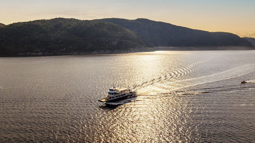 Un ferry navigue sur une étendue d'eau paisible sous un coucher de soleil doré. Le ciel est partiellement nuageux, et des collines recouvertes d'une dense forêt verte encadrent la scène en arrière-plan. Le ferry laisse derrière lui un sillage doux qui se dissipe lentement.