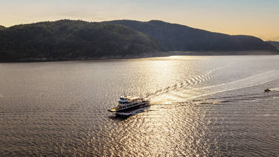 A ferry sails across a tranquil body of water under a golden sunset. The sky is partly cloudy, and hills covered with dense green forest frame the scene in the background. The ferry leaves a gentle wake behind.