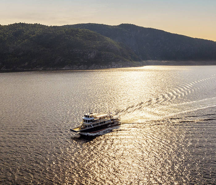 A ferry sails across a tranquil body of water under a golden sunset. The sky is partly cloudy, and hills covered with dense green forest frame the scene in the background. The ferry leaves a gentle wake behind.