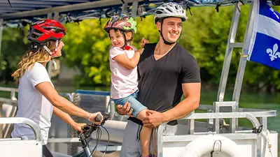 Une famille souriante portant des casques de vélo est à bord d'un bateau, avec un vélo et un drapeau du Québec visible à l'arrière-plan.