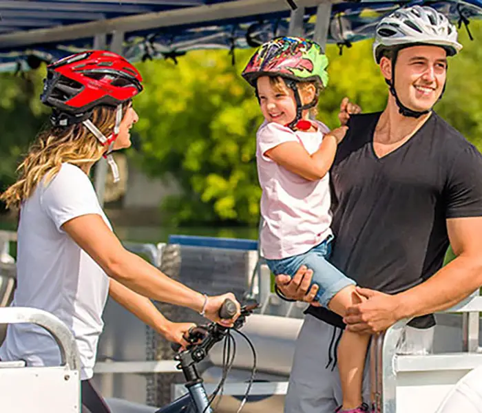 Une famille souriante portant des casques de vélo est à bord d'un bateau, avec un vélo et un drapeau du Québec visible à l'arrière-plan.