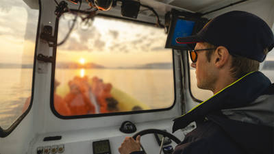 A captain wearing a cap and sunglasses steers a boat while looking through the windshield. Outside, a sunset over the water is visible, with blurred passengers at the back of the boat. The sky is partly cloudy, creating a warm atmosphere.