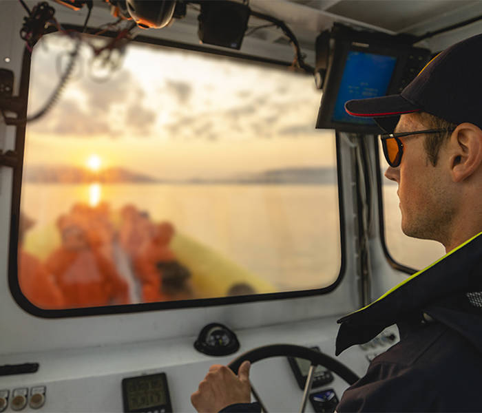 A captain wearing a cap and sunglasses steers a boat while looking through the windshield. Outside, a sunset over the water is visible, with blurred passengers at the back of the boat. The sky is partly cloudy, creating a warm atmosphere.