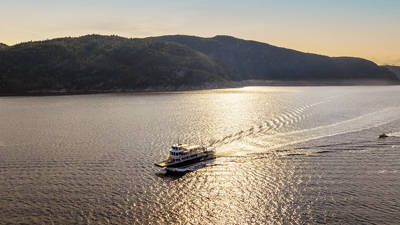 A cruise ship sails on a river at sunset, leaving a trail in the shimmering water. In the background, green hills line the river under a golden, peaceful sky.