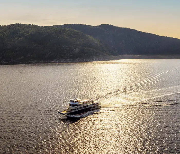 Un bateau de croisière navigue sur une rivière au coucher du soleil, laissant une traînée dans l'eau scintillante. À l'arrière-plan, des collines verdoyantes bordent la rivière sous un ciel doré et paisible.