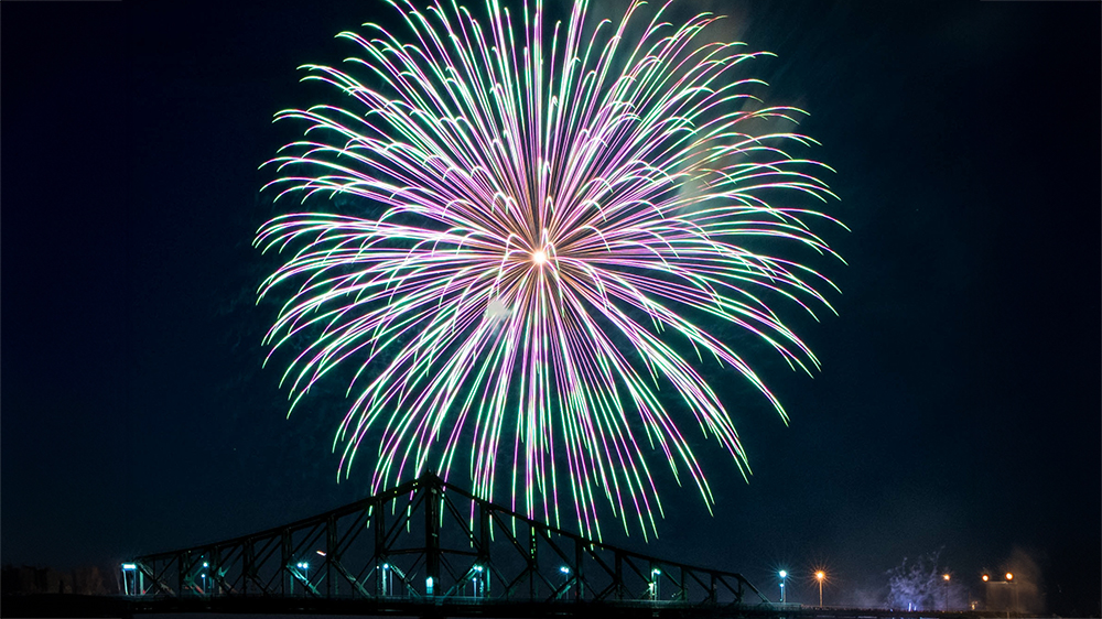 A massive multicolored firework bursts in the night sky above an illuminated bridge, creating a spectacular and vibrant scene.