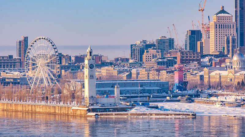 Vue de Montréal en hiver depuis le Vieux-Port avec la Tour de l'Horloge au premier plan, puis la grande roue derrière, et la skyline de Montréal enneigée en arrière plan.