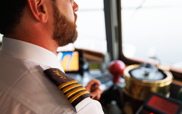 Un capitaine de navire en uniforme, vu de dos, surplombe l'équipement de navigation et le fleuve Saint-Laurent depuis le pont d'un navire.