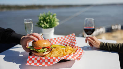 A plate with a hamburger, chips, and a pickle on a white table with a red-and-white checkered pattern. One person holds a glass of red wine, another touches the burger. A river and a potted plant are visible in the background.