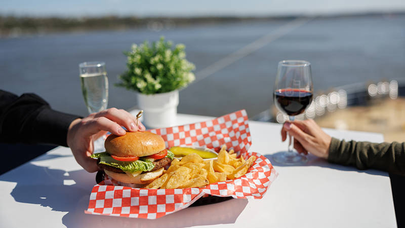 A plate with a hamburger, chips, and a pickle on a white table with a red-and-white checkered pattern. One person holds a glass of red wine, another touches the burger. A river and a potted plant are visible in the background.