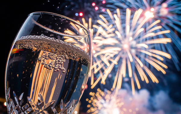 A close-up of a sparkling champagne flute in the foreground, with vivid fireworks lighting up the night sky in the background.