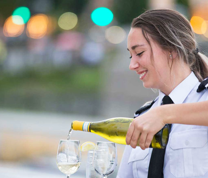 Une serveuse souriante servant du vin blanc dans deux verres posées sur une table. 