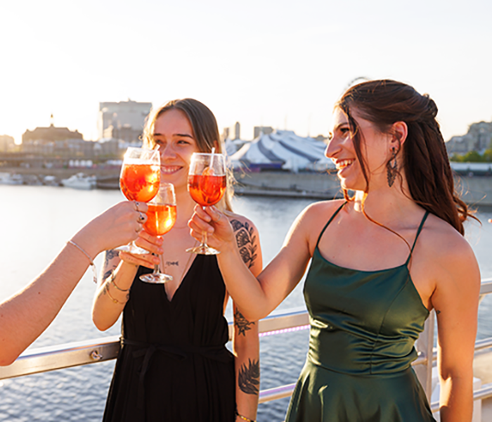 Three women dressed in elegant attire toasting with cocktails on the terrace of the AML Cavalier Maxim ship in Montreal