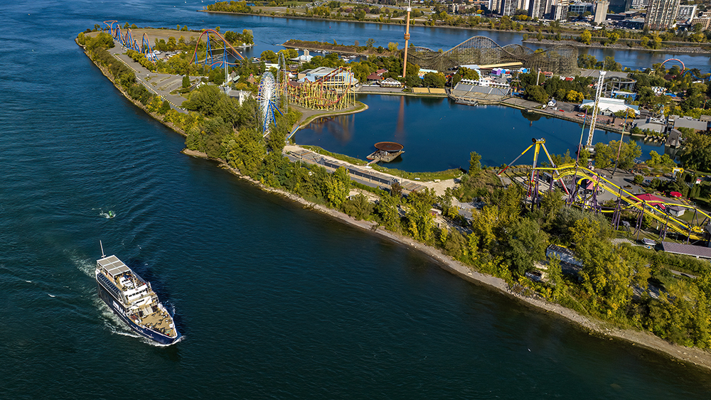 Aerial view of an island with an amusement park featuring roller coasters, a Ferris wheel, and rides, surrounded by a river. A cruise ship is sailing on the water to the left.