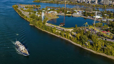 Aerial view of an island with an amusement park featuring roller coasters, a Ferris wheel, and rides, surrounded by a river. A cruise ship is sailing on the water to the left.