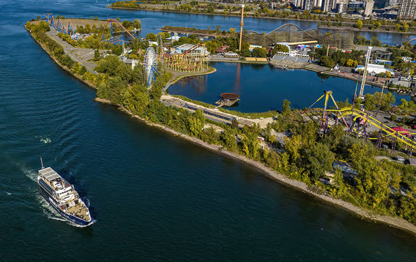 Aerial view of an island with an amusement park featuring roller coasters, a Ferris wheel, and rides, surrounded by a river. A cruise ship is sailing on the water to the left.