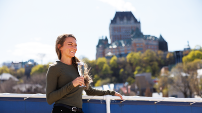 A smiling woman holding a Champagne flute on one of the terraces of the AML Louis Jolliet ship in Quebec City, with the Château Frontenac in the background.