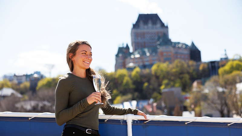 Une femme souriante tenant une flûte de Champagne sur l'une des terrasses du navire AML Louis Jolliet à Québec avec en arrière plan le Château Frontenac.