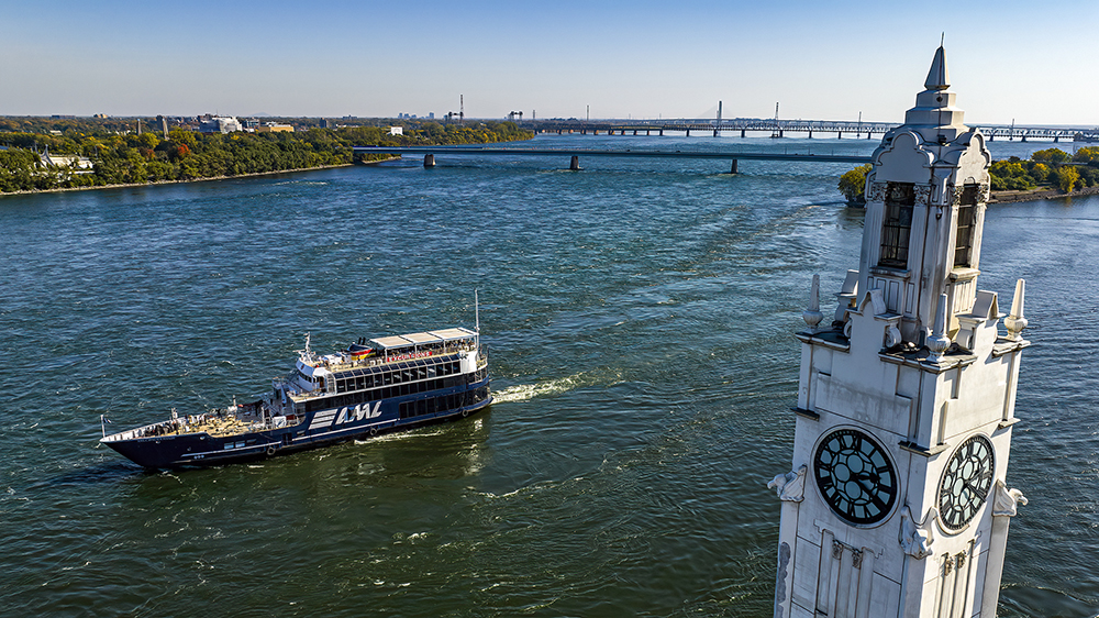 Aerial view of a cruise ship sailing on a river, with a large white clock tower in the foreground and a bridge in the background under a clear sky.