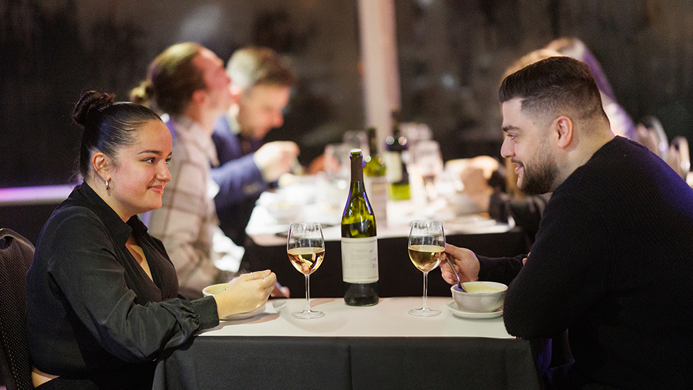 A couple shares a meal in an elegant setting, with two glasses of white wine and a bottle on the table. Other diners are visible in the background.