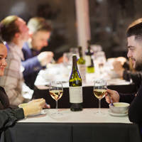 Un couple partage un repas dans une ambiance élégante, avec deux verres de vin blanc et une bouteille sur la table. D'autres convives sont visibles en arrière-plan.