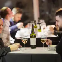 A couple shares a meal in an elegant setting, with two glasses of white wine and a bottle on the table. Other diners are visible in the background.