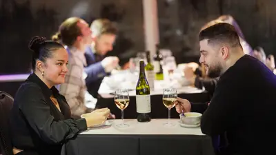 A couple shares a meal in an elegant setting, with two glasses of white wine and a bottle on the table. Other diners are visible in the background.