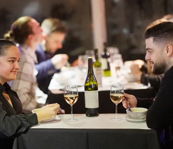 A couple shares a meal in an elegant setting, with two glasses of white wine and a bottle on the table. Other diners are visible in the background.