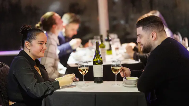 A couple shares a meal in an elegant setting, with two glasses of white wine and a bottle on the table. Other diners are visible in the background.