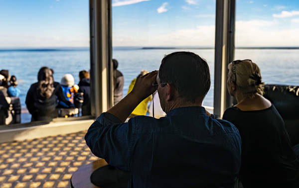 A group of people relax on a boat, viewing a vast, calm ocean. A man in the foreground uses binoculars, while a woman sits beside him. The scene is sunny and serene, with clear blue skies and distant horizons.